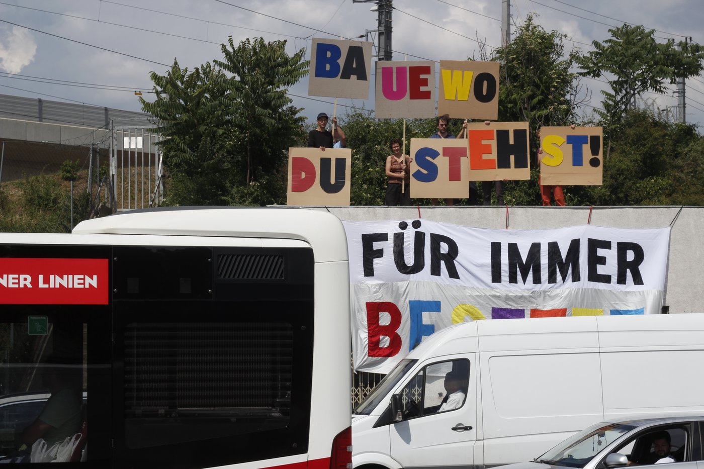 im vordergrund sieht man einen auschnitt einer verkehrssituation mit autos im stau – im hintergrund ein gehisstest transparent und dahinter 4 personen mit demonstrierten schildern, auf denen in bunten buchstaben „baue wo du stehst“ steht.