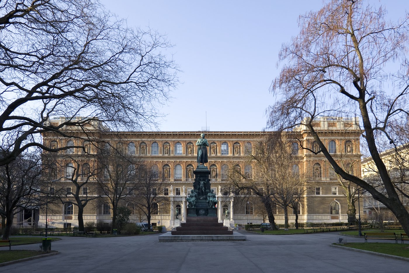 3a Im Zentrum des Fotos befindet sich die Vorderfront des Gebäudes der Akademie der bildenden Künste Wien. Das viergeschossige Gebäude hat viele hohe halbrunde Fenster, zwischen denen sich halbrunde Nischen mit Skulpturen befinden. Die ockerfarbene Fassade ist reich gestaltet und hat einen weißen Sockel. Das Dach ist flach und die Vorderfont ist in einen mittleren breiten und zwei turmähnliche Seitenteile gegliedert. Vor dem Gebäude befindet sich der Schillerplatz mit dem Park und dem Schillerdenkmal. Da die Bäume ganz kahl sind, kann davon ausgegangen werden, dass das Foto im Winter gemacht wurde.