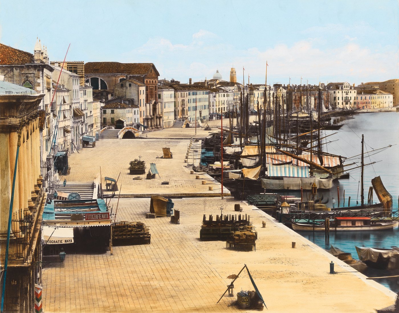 Naya Koloriertes Foto der Uferpromenade Riva degli Schiavoni in Venedig, die von einer Häuserreihe links und der Lagune mit Segelschiffen rechts gesäumt wird. Links im Vordergrund ist das Geschäft des Fotografen zu erkennen.