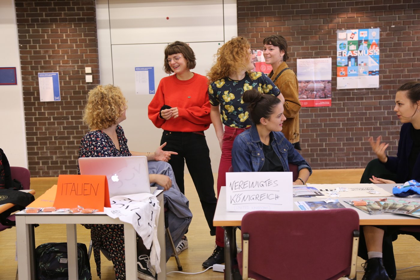 Exchange Fair des International Office, 2018, Aula in der Augasse, Foto © Iklim Dogan Man sieht Incoming Austauschstudierendeaus Italien und dem vereinigten Königreich im lebhaften Gespräch mit Studierenden, die sich für einen Auslandsaufenthalt interessieren. An der Rückwand hängt ein Plakat, welches auf das Mobilitätsprogramm Erasmus plus aufmerksam macht.