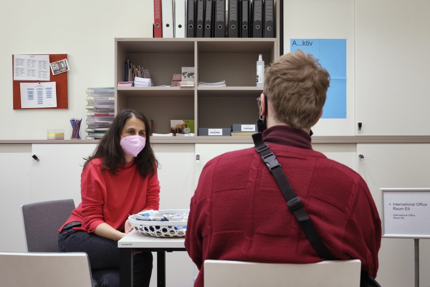 Beratungsgespräch im International Office, Foto © Thomas Freiler Eine Frau und ein Student sitzen an einem Tisch, beide haben rote Pullover und Masken an. Im Hintergrund sieht man typische Büroeinrichtung.