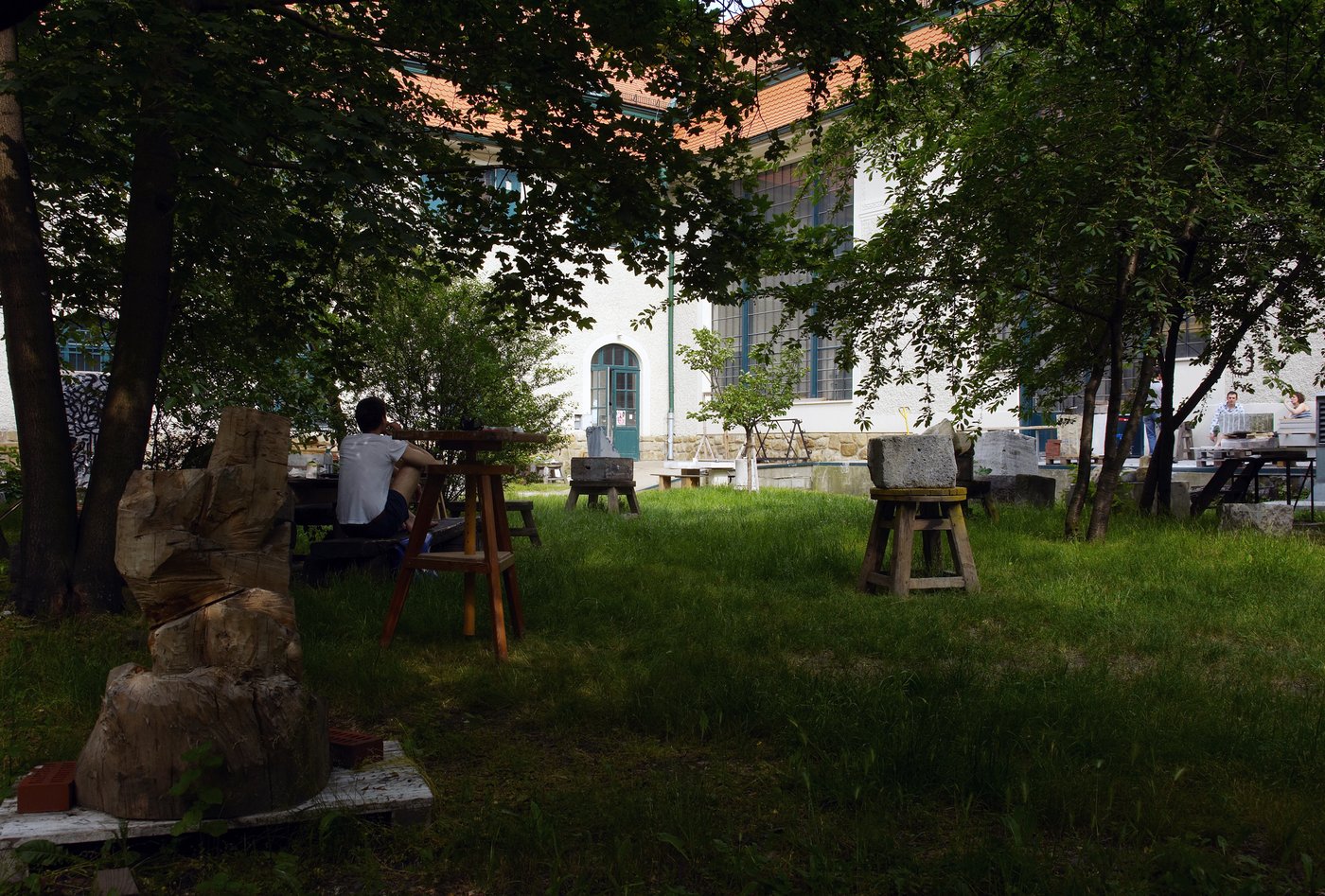 Blick in den Garten der Bildhauereiateliers, Foto © Lisa Rastl In einem sommerlichen Garten sitzt ein junger Mann, den Rücken zugewandt, die Beine angezogen, mit einem weißen T-Shirt. Er ist umgeben von Holz- und Steinskulpturen. Im Hintergrund sieht man das historische Gebäude mit den großen Atelierfenstern.